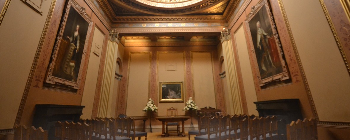 a peach coloured room with rows of chairs a desk and a large fireplace